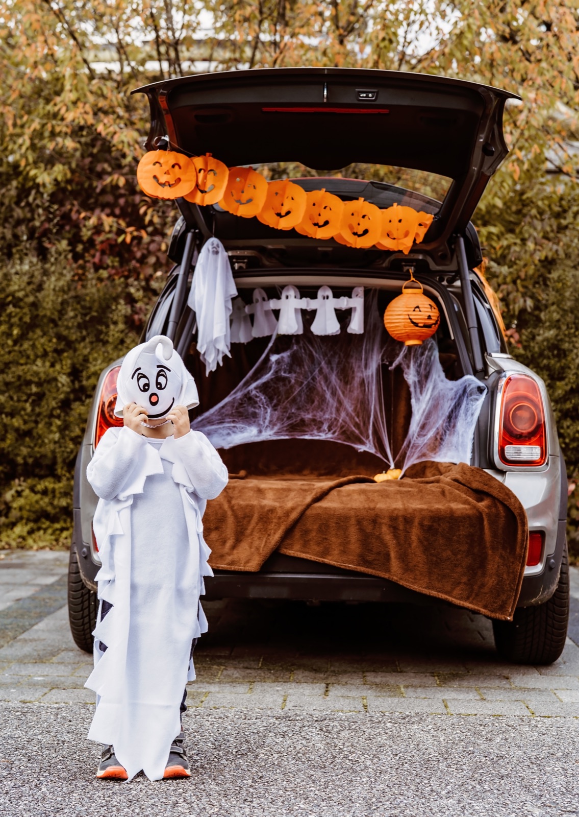 A child dressed as a friendly ghost standing beside a Halloween-decorated car trunk